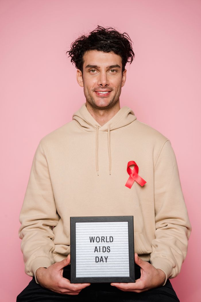 Smiling man holding sign with World AIDS Day message and red ribbon on pink background.
