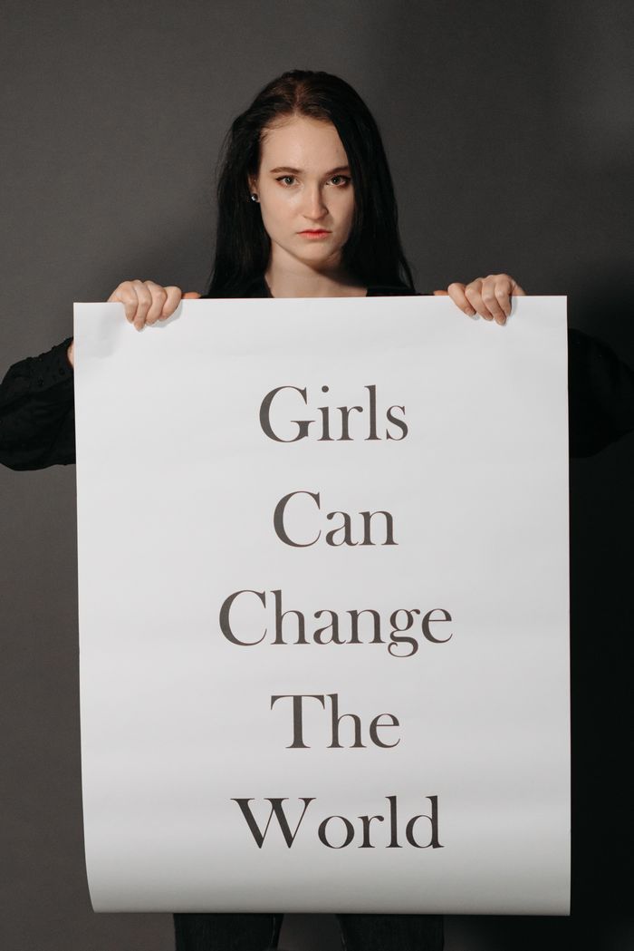 A woman holds a powerful sign stating 'Girls Can Change the World' on a gray background.